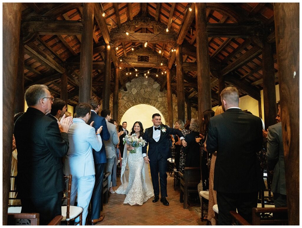 Savana and Casey walk back down the aisle hand-in-hand after their first kiss, beaming with happiness as their guests applaud inside Ian's Chapel.