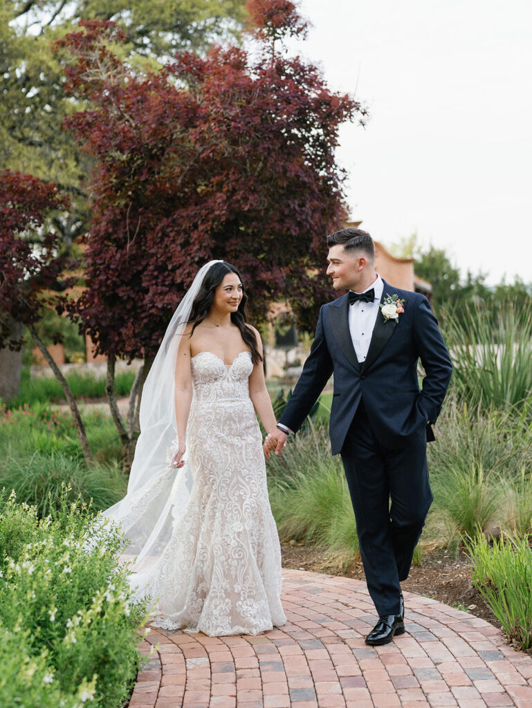 Casey gently holds Savana's hand as they walk along a flower-lined path, with Ian's Chapel visible in the distance at Camp Lucy.