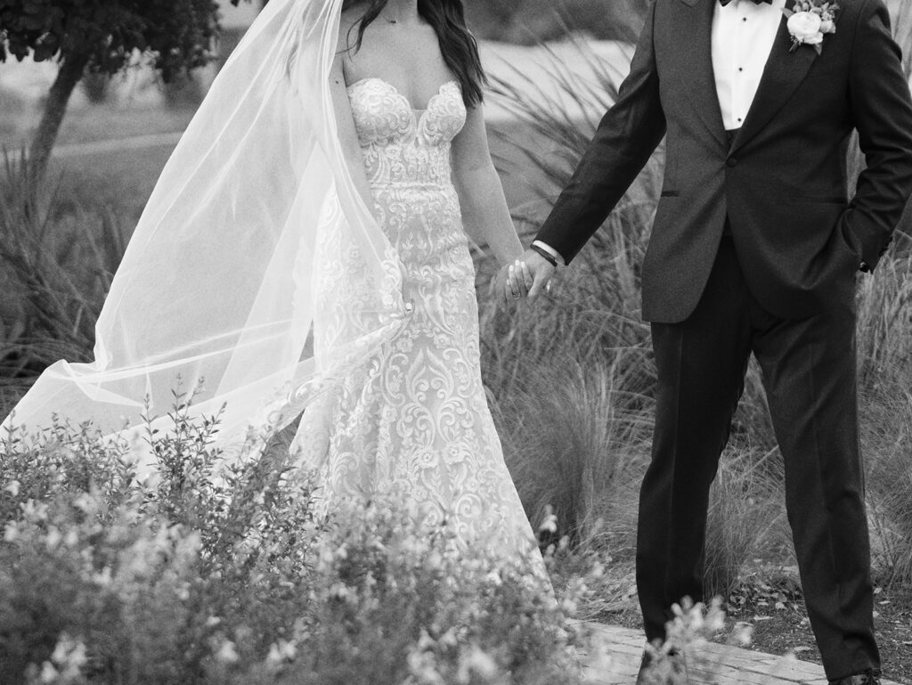 Casey gently holds Savana's hand as they walk along a flower-lined path, with Ian's Chapel visible in the distance at Camp Lucy.